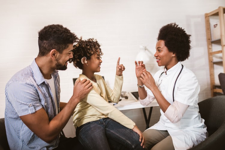 A smiling doctor in a white coat sits at a desk signing with a pediatric patient.