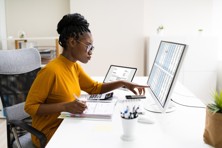 A person at a desk takes notes in a binder while looking at data on a computer.