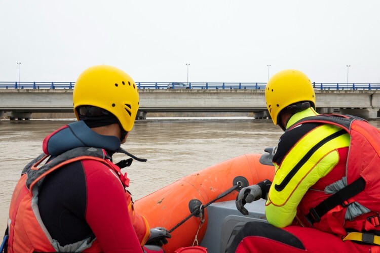 Two emergency rescuers survey floodwater from a boat.