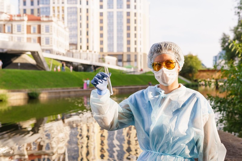 An epidemiologist in a mask, gloves, surgical cap, and gown stands outside a community civic center holding up a test tube.