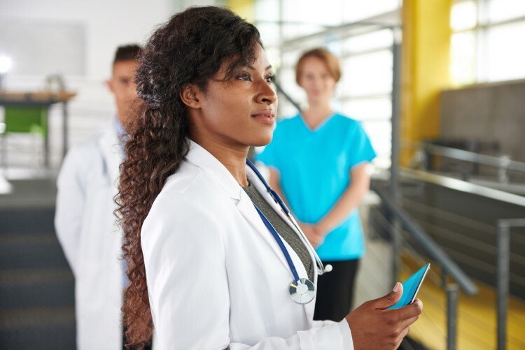 A healthcare manager in a lab coat holding a smartphone stands in a hospital room with two colleagues.
