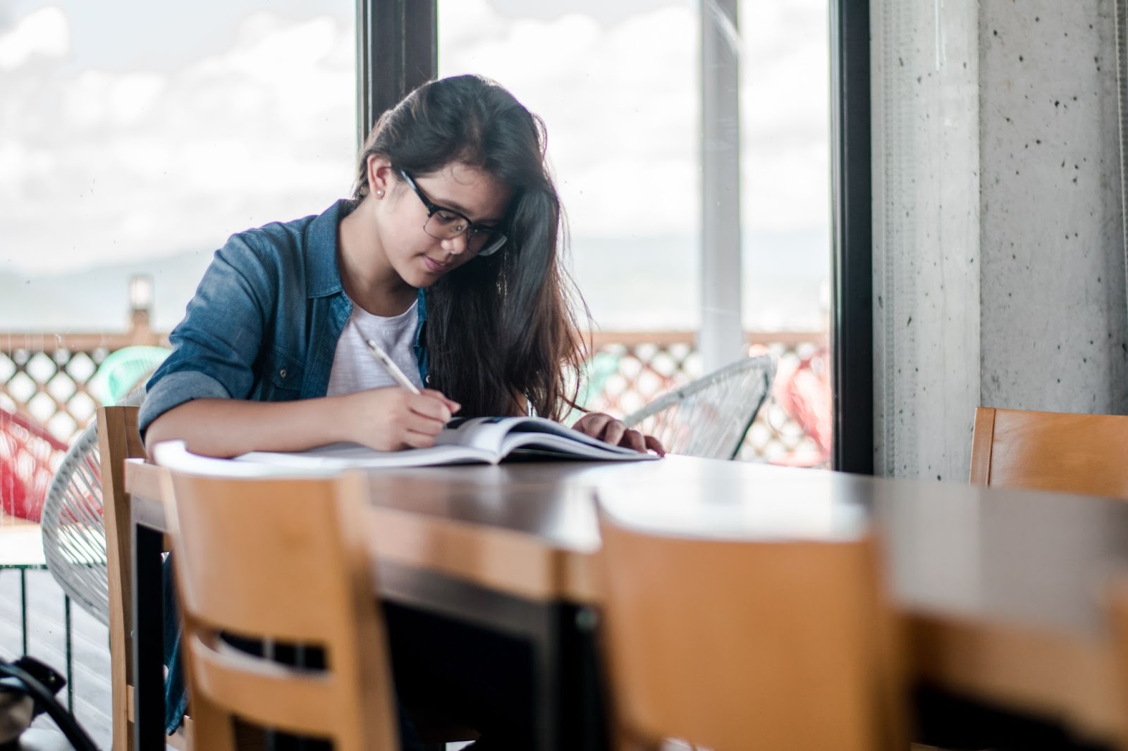 A female student with glasses writes in a book while sitting at a table.