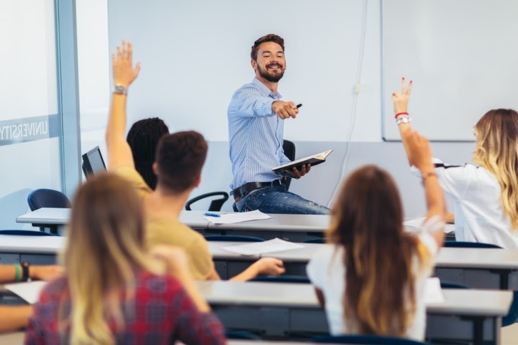 A teacher smiles and points to a group of high school-age students sitting at classroom desks who are raising their hands to be called on.