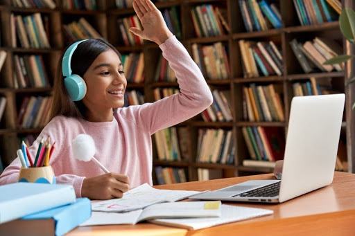 A student wearing headphones raises their hand and smiles at a laptop screen during virtual instruction
