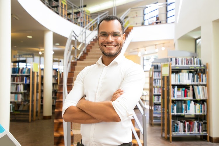 A smiling teacher wearing glasses stands in a school library.