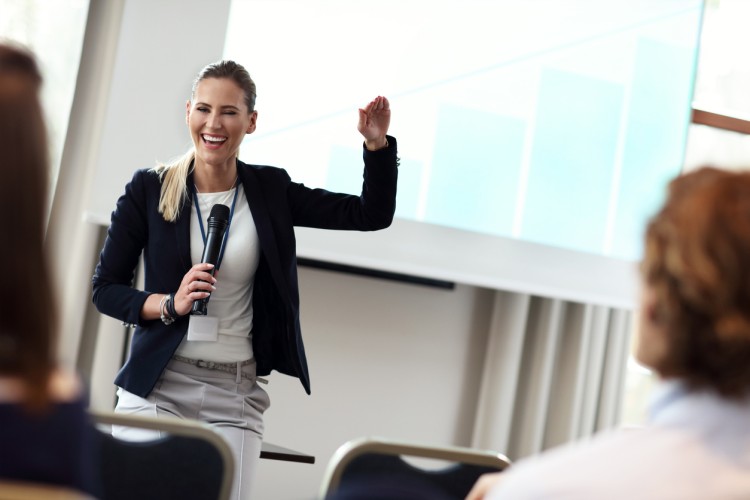 A person in business attire holds a microphone while speaking to a group.