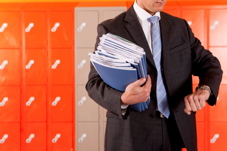Against a background of school lockers, a district administrator in a suit and tie and carrying a stack of folders checks the time on his wristwatch.