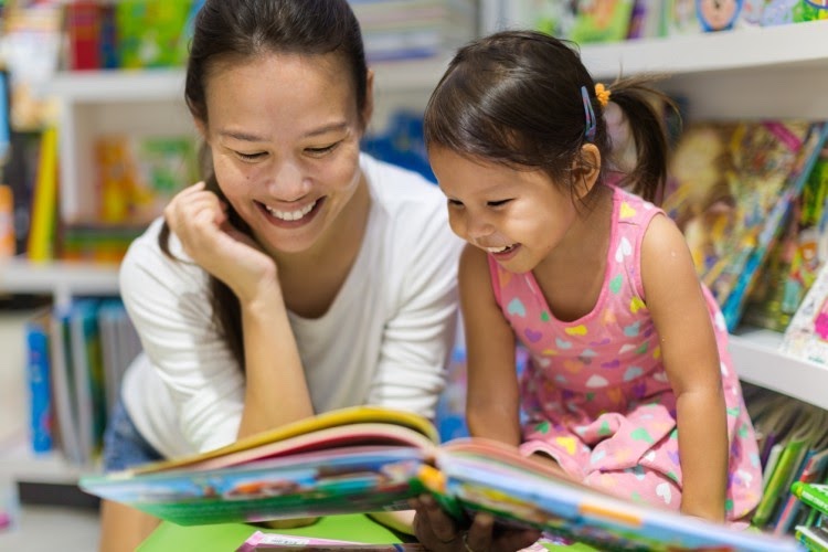 A mother and her daughter read a picture book together in a library.