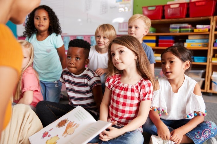 A diverse group of kindergarten students sits on the rug in their classroom while their teacher reads them a picture book.