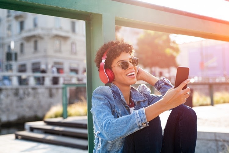 Woman in sunglasses wears headphones and looks at phone.