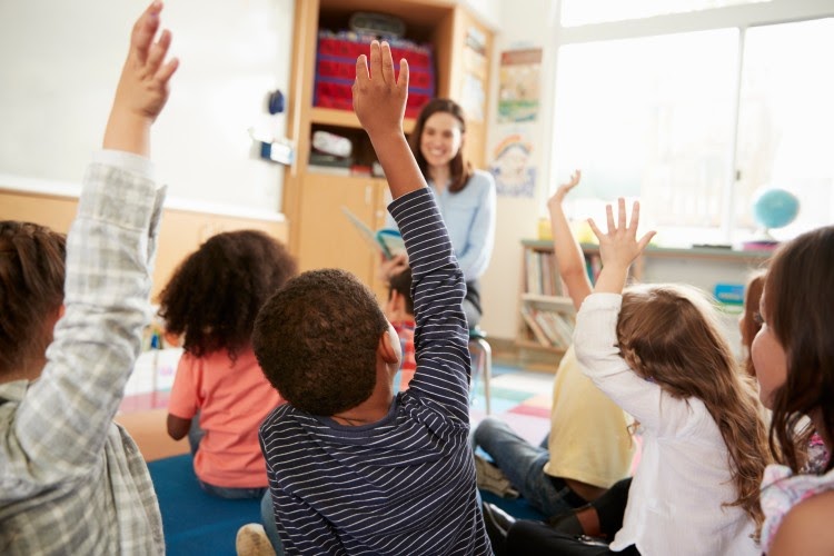 Children in classroom sit on floor raising hands.