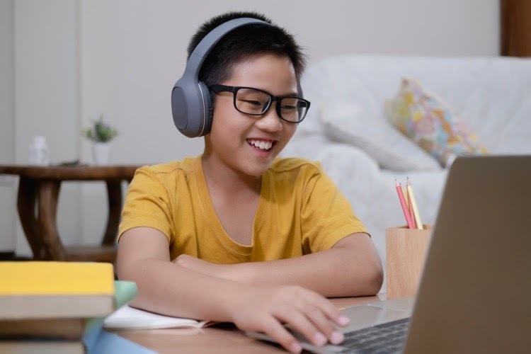 A young person at home wearing headphones smiles while watching a laptop screen during a lesson.