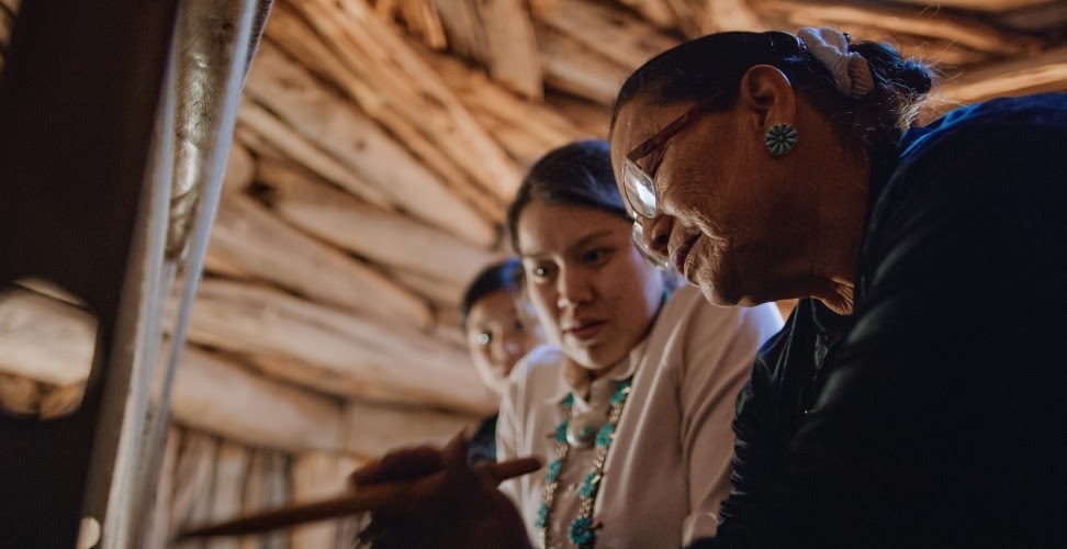 A native american woman teaches how to weave at a loom.