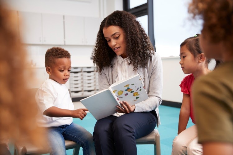 A teacher holds a book and reads to a group of young students.