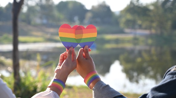 Close-up of two people’s hands, each raising a rainbow heart against a background of a lake and trees.