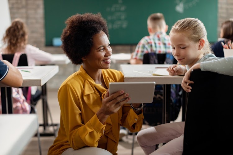 A teacher holding a tablet kneels by a student sitting at their desk.