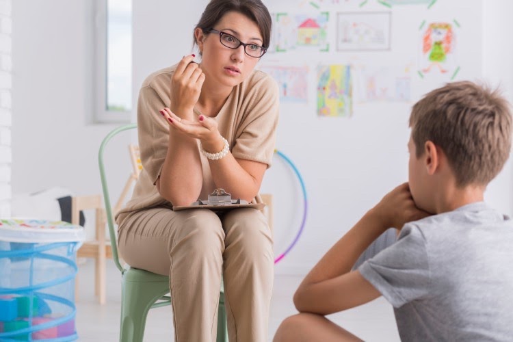 A teacher sits in a chair in an elementary school classroom talking to a little boy seated on the floor.