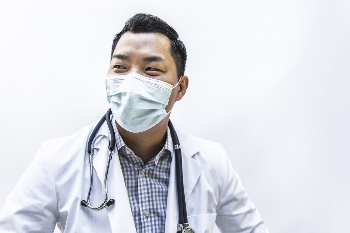 A male healthcare professional wearing a white lab coat and face mask looks away from the camera.
