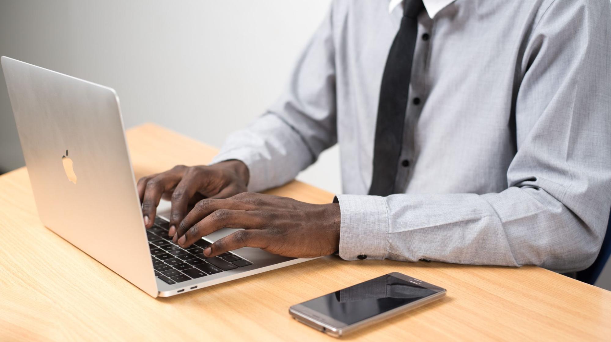A man types on a laptop next to a cell phone.