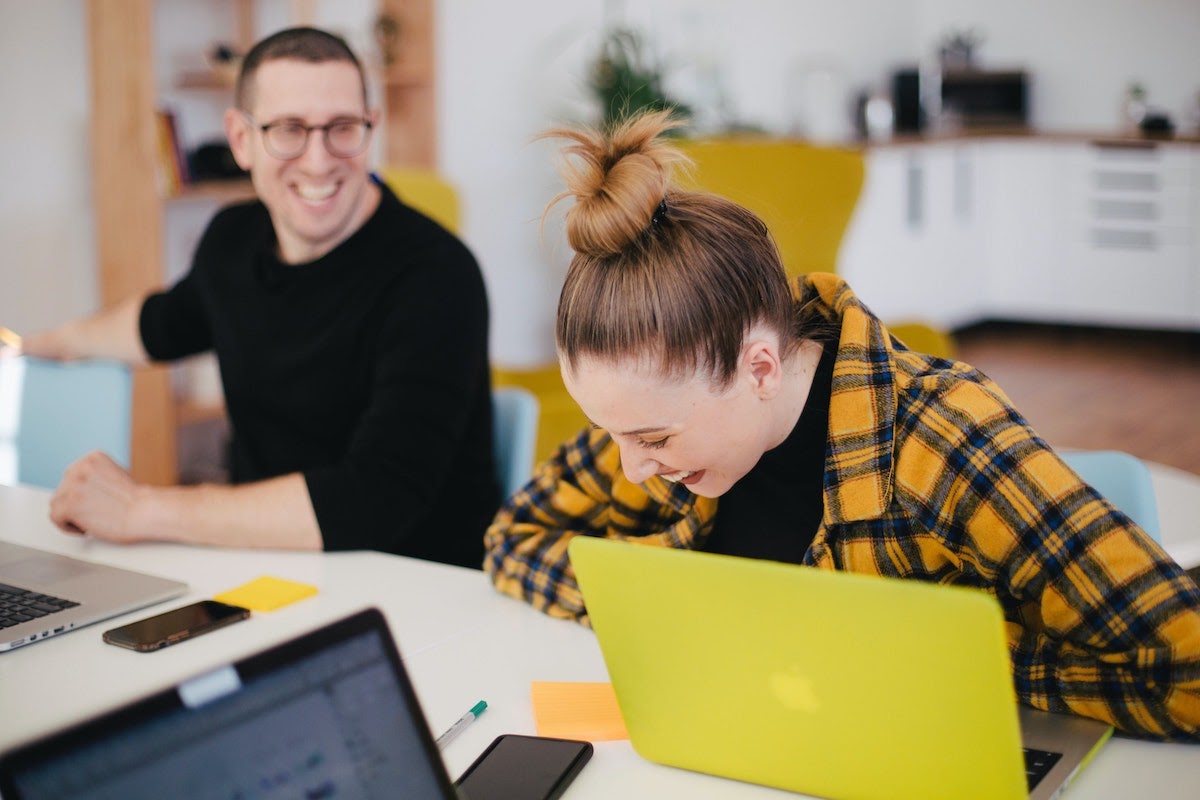A man and woman laugh while sitting in front of their laptops.